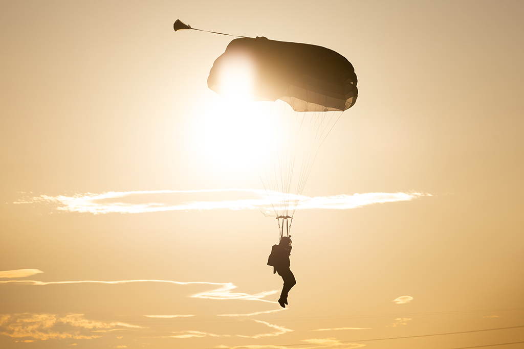 Silhouette of a lone Royal Air Force Parachuting Test Team member descending under an open parachute, backlit by a bright, glowing sun in a golden hazy sky.